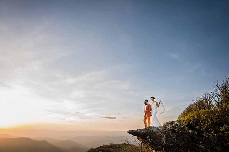 A couple in wedding attire stands on a rocky cliff edge at sunset, where an Event Audio Company ensured every vow echoes perfectly. The bride wears a white dress and veil, the groom in a pink suit. Mountains stretch beneath a sky dotted with clouds, warmly lit by the sun from the left.