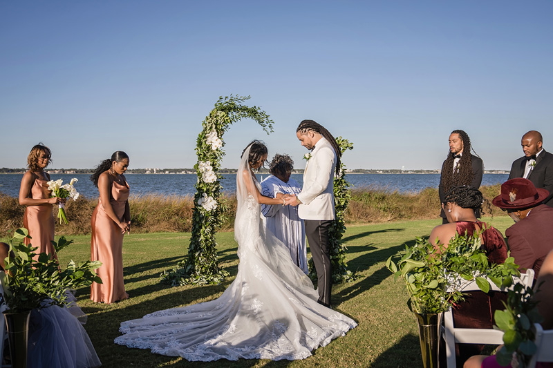 A couple stands under a floral arch during an outdoor wedding ceremony by a lake, where the Event Audio Company ensures perfect sound. The bride wears a long white gown with a train, and the groom wears a white jacket and black pants. Bridesmaids in pink dresses and groomsmen in suits stand nearby as guests are seated, witnessing the event.