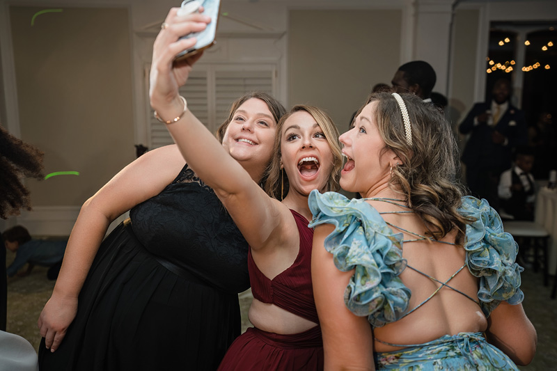 Three women happily take a selfie together at an Event Audio Company gathering. The woman on the left wears a black dress, the woman in the middle sports a red dress, and the woman on the right dazzles in a blue dress with ruffled sleeves. They're indoors, with soft lighting and people in the background.