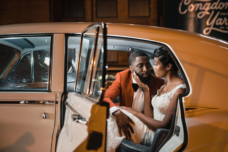 A bride and groom sit inside a classic car with the door open. The groom, in a rust-colored suit, gently holds the bride's chin as she gazes at him in her lace wedding gown. "Congdon Yards" glows softly in the background, setting the perfect scene orchestrated by an Event Audio Company.