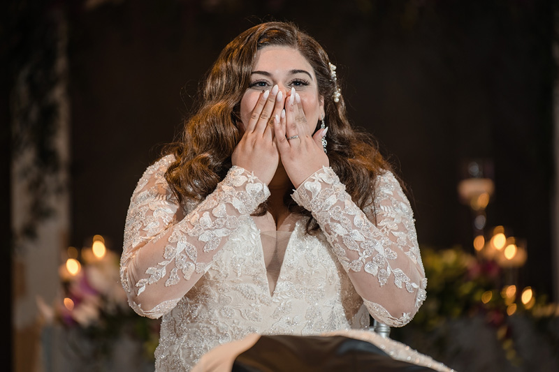 A bride with long brown hair and a lace wedding dress covers her mouth in surprise or excitement, the ambient glow of soft candles and flowers enhanced by the harmonious touch of an Event Audio Company. Her floral hair accessory complements the warm, romantic atmosphere beautifully.