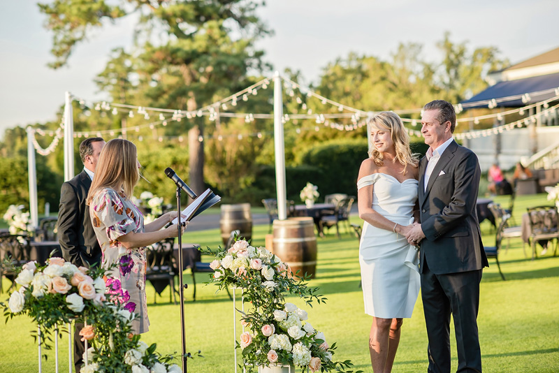 A couple stands in an outdoor setting, surrounded by lush greenery and string lights. The woman wears a white dress, and the man is in a dark suit. As a person from the event audio company speaks at a microphone, floral arrangements with pink and white flowers embellish the scene. Tables and chairs are in the background.