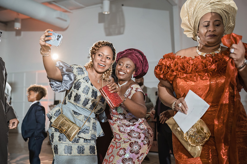 Three women in vibrant, traditional African attire take a selfie indoors at an event organized by an Event Audio Company. The leftmost wears silver with gold accessories, the center sports a burgundy headwrap and floral dress, and the rightmost dazzles in orange with a matching gele. A child is visible in the background.