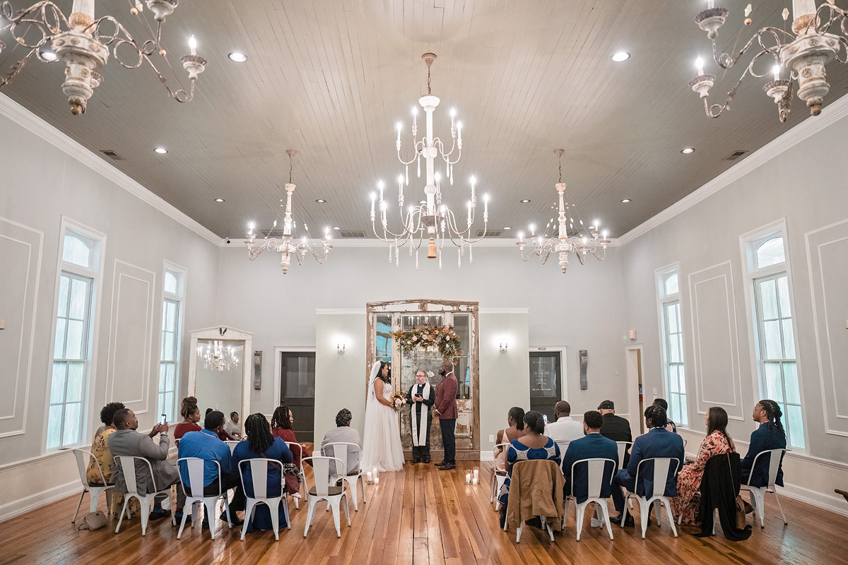 A wedding ceremony unfolds in a charming, high-ceilinged room with wooden floors and elegant chandeliers. The couple stands before a floral arch, while guests seated in two rows listen intently, thanks to the Event Audio Company ensuring crystal-clear sound. Natural light pours through large windows lining the side walls.