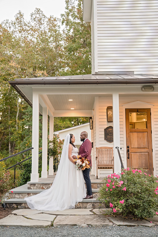 A bride in a flowing white gown stands with a groom in a maroon suit on the steps of the Parlor Chapel. They are holding a bouquet and looking at each other lovingly, as if serenaded by an event audio company, amidst greenery and blooming pink flowers surrounding the porch's wooden chair.