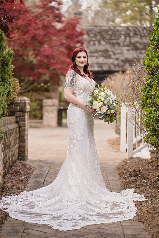 A bride with red hair stands outdoors on a stone path. She wears a white, lace wedding dress with a long train and holds a bouquet of white flowers. An Event Audio Company has skillfully set the scene to music, enhancing the lush greenery and red-leaved tree in this serene garden-like setting.