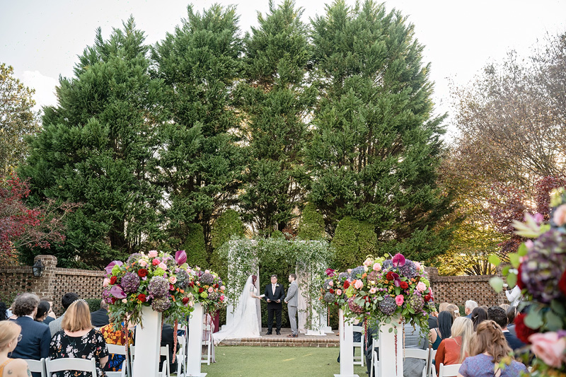 A wedding ceremony unfolds outdoors against a lush backdrop of tall green trees at The Sutherland. The couple stands with an officiant under a floral arch, their vows beautifully amplified by an Event Audio Company. Guests sit on either side, colorful flower arrangements lining the aisle in the bright, festive atmosphere.