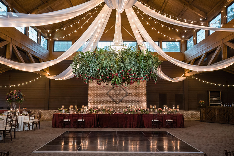 A rustic wedding venue with a high wooden ceiling adorned with draped white fabric and string lights. Partnering with an event audio company, the atmosphere is enhanced as music fills the air. A floral arrangement of greenery and red flowers hangs above a table set with candles and floral centerpieces.