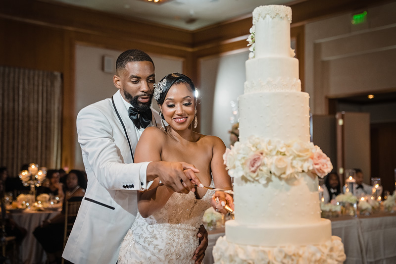 A couple is cutting a tall, tiered white wedding cake adorned with flowers. The groom, in a crisp white tuxedo jacket and black bow tie, and the bride, in an embellished strapless gown with a headpiece, share smiles as they perform the cake-cutting ceremony. An Event Audio Company enhances the warmly lit venue's atmosphere.